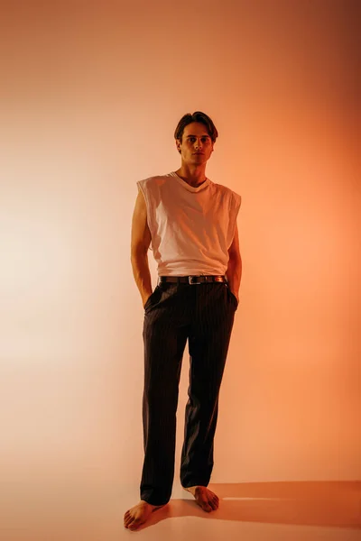 Full length of stylish man in white t-shirt and black pants posing with hands in pockets on orange — Stock Photo