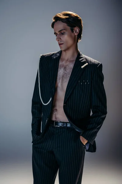 Morena hombre con el pecho peludo y elegante chaqueta con cadena de perlas posando con las manos en bolsillos en gris oscuro - foto de stock