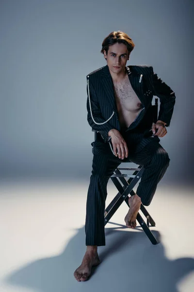 Повна довжина стильного чоловіка з волохатими грудьми і модний костюм з перловим ланцюгом, що сидить на кріслі табору на темно-сірому — стокове фото