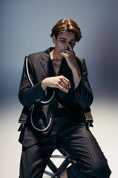 Hombre elegante en traje de moda con cadena de perlas sentado en la silla del campamento mientras mira a la cámara en gris oscuro - foto de stock