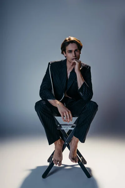 Full length of barefoot man in trendy suit with pearl chain sitting on camp chair while looking at camera on grey — Stock Photo