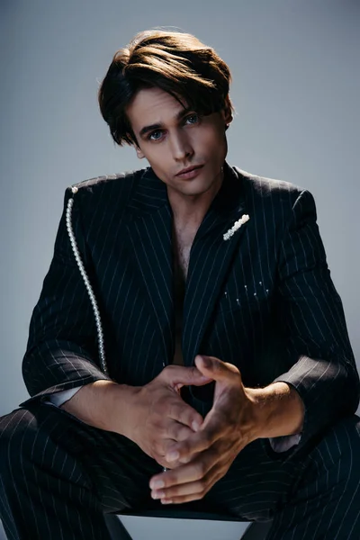 Stylish man in trendy suit with pearl chain sitting while looking at camera on dark grey — Stock Photo