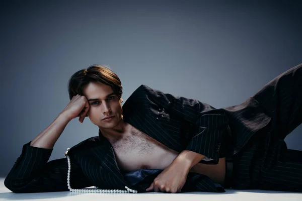 Hombre elegante en chaqueta con perlas y pecho peludo posando sobre fondo gris oscuro - foto de stock