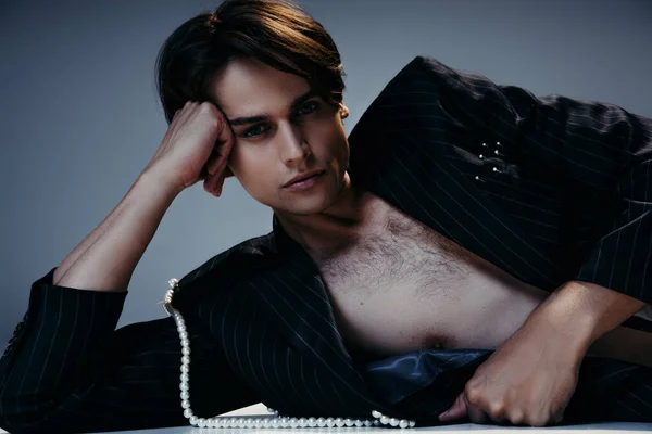 Elegante hombre de chaqueta con perlas y pecho peludo posando sobre fondo gris oscuro - foto de stock