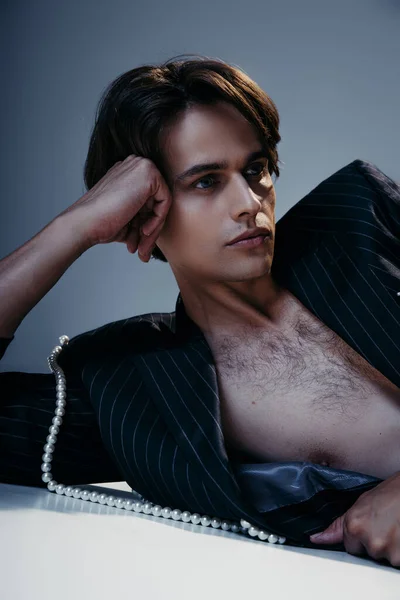 Elegante hombre de chaqueta con perlas y pecho peludo mirando hacia otro lado sobre fondo gris oscuro - foto de stock