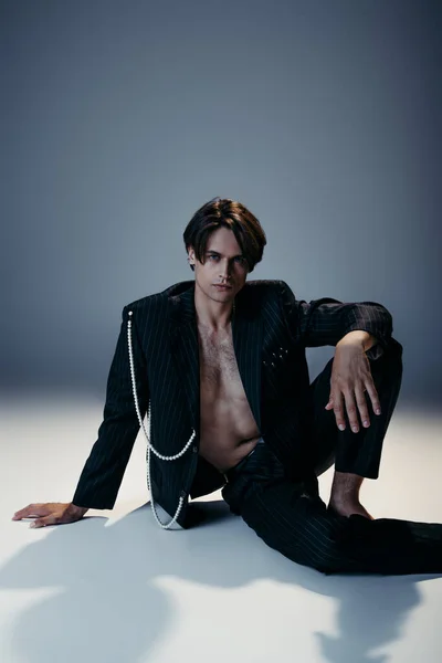 Stilvoller Mann in trendiger Jacke mit Perlen und behaarter Brust sitzt auf dunkelgrauem Hintergrund — Stockfoto