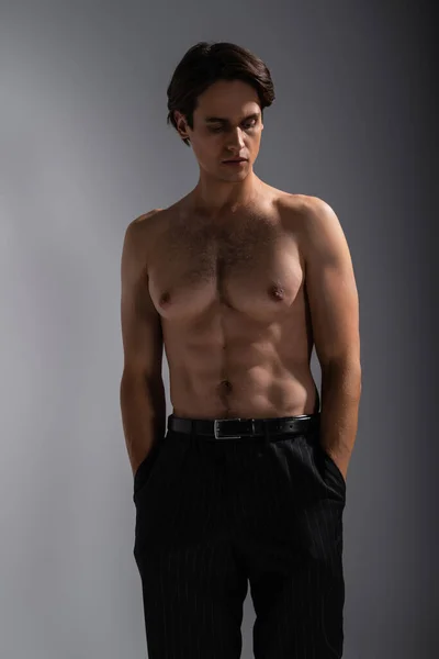 Gut aussehender Mann mit muskulösem Körper posiert mit Händen in Taschen schwarzer Hosen auf grau — Stockfoto