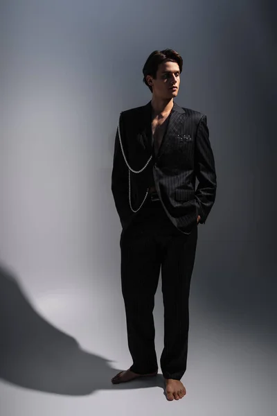 Full length of man in trendy suit with pearls looking away while posing with hands in pockets on dark grey — Stock Photo