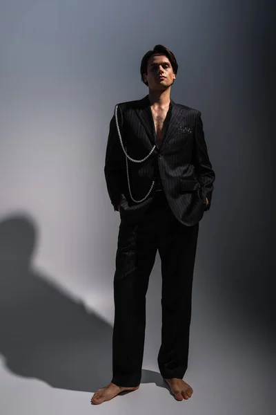 Barfuß in voller Länge Mann im trendigen Anzug mit Perlen posiert mit Händen in Taschen auf dunkelgrau — Stockfoto