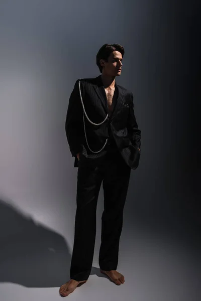 Повнометражний босоніж чоловік у модному костюмі з перлами, дивлячись далеко, позуючи руками в кишенях на темно-сірому — стокове фото