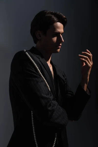 Uomo in giacca alla moda con perle gesticolanti mentre posa su grigio scuro — Foto stock