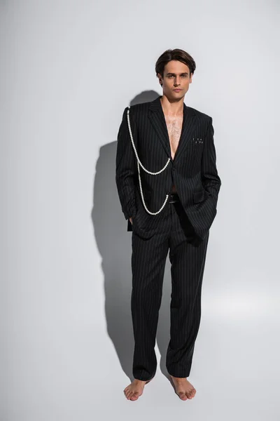 Full length of barefoot man in trendy suit with pearls posing with hands in pockets on grey — Stock Photo