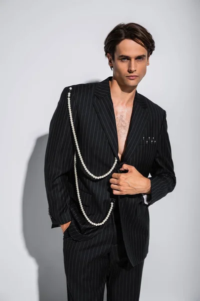 Morena hombre en chaqueta de moda con perlas posando mientras mira a la cámara en gris - foto de stock