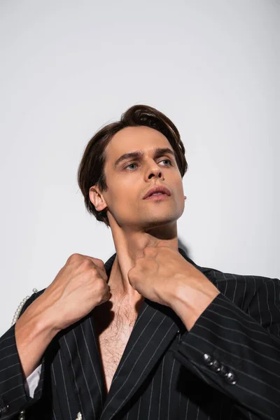 Morena hombre con ojos azules ajustando chaqueta de moda con perlas aisladas en gris - foto de stock