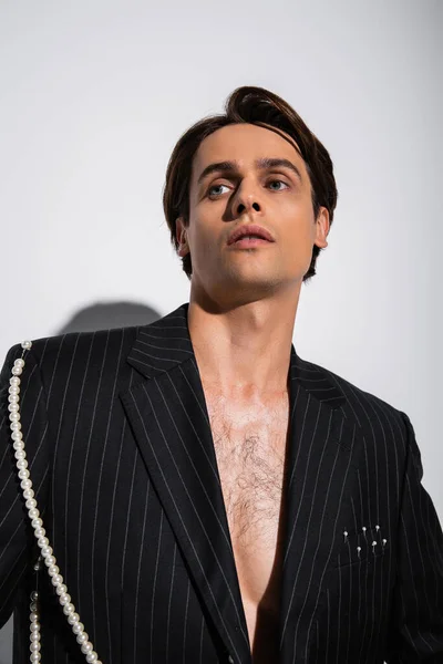 Bruna uomo in giacca alla moda con strisce e perle in posa mentre distoglie lo sguardo sul grigio — Foto stock