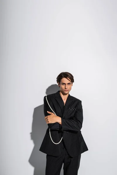 Morena hombre en blazer de moda con perlas posando con los brazos cruzados mientras mira a la cámara sobre fondo gris - foto de stock