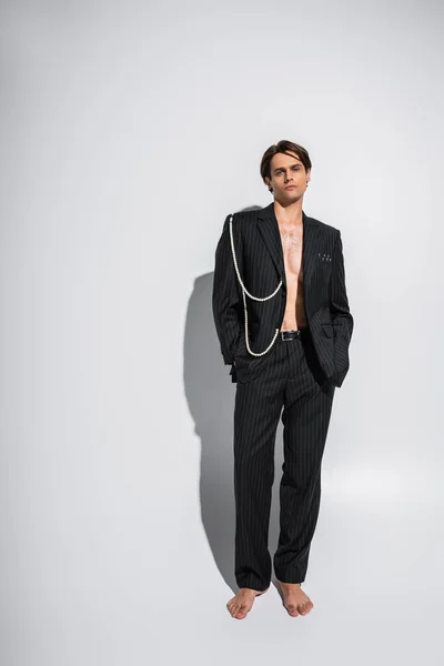 Full length of barefoot and brunette man in trendy suit with pearls posing with hands in pockets on grey — Stock Photo