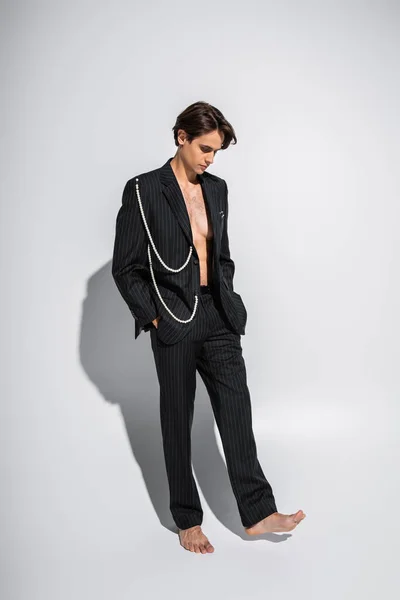 Full length of barefoot and brunette man in trendy suit with pearls standing with hands in pockets on grey — Stock Photo