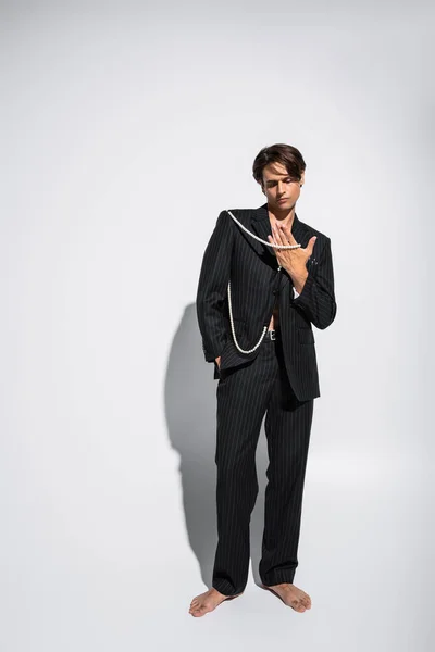 Full length of barefoot and brunette man in trendy suit holding pearls while posing with hand in pocket on grey — Stock Photo