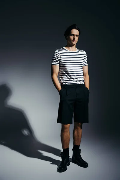 Повна довжина модного чоловіка в смугастій футболці і шортах тримає руки в кишенях на сірому фоні з освітленням — стокове фото