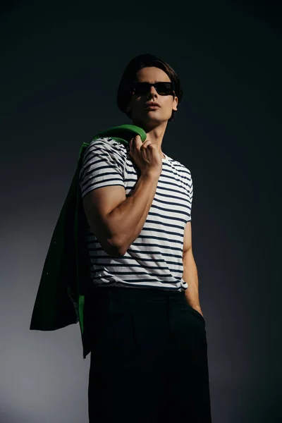Vue à angle bas de l'homme à la mode en lunettes de soleil posant avec veste en cuir vert sur fond gris avec éclairage — Photo de stock