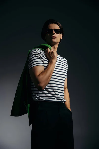 Low angle view of trendy man in sunglasses posing with green leather jacket on grey background with lighting — Stock Photo