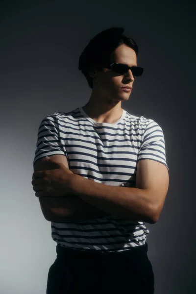 Trendy model in striped t-shirt and sunglasses crossing arms on grey background with lighting — Stock Photo