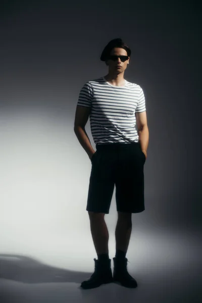 Full length of trendy man in sunglasses holding hands in pockets of shorts on grey background with lighting — Stock Photo