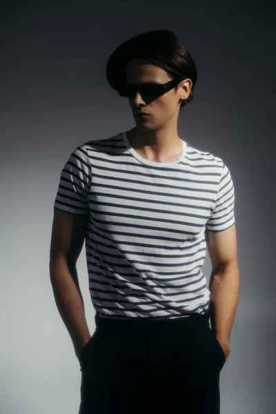 Стильний чоловік в береті і сонцезахисних окулярах тримає руки в кишенях шортів на сірому фоні з освітленням — стокове фото