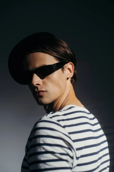 Portrait of brunette man in stylish sunglasses and striped t-shirt standing on grey background with lighting — Stock Photo