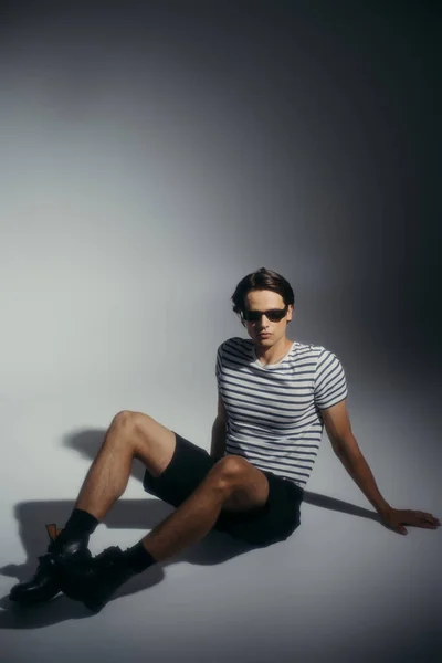 Homme branché en lunettes de soleil et chaussures assis sur fond gris avec éclairage et ombre — Photo de stock