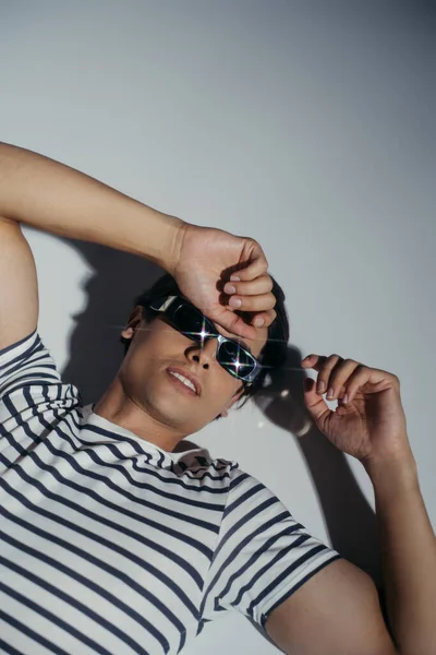 Top view of smiling man in sunglasses and striped t-shirt lying on grey background with shadow — Stock Photo