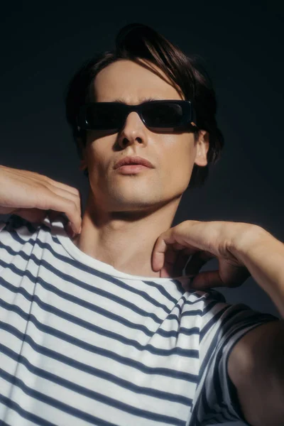 Hombre de moda en gafas de sol tocando camiseta rayada aislada en gris con iluminación - foto de stock