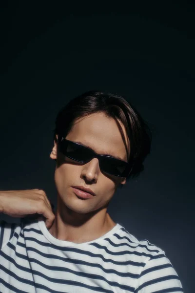 Modelo morena de moda en gafas de sol tocando camiseta aislada en gris con iluminación - foto de stock