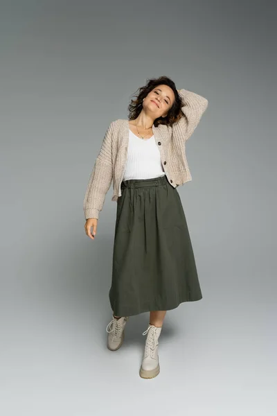 Full length of trendy woman in knitted cardigan and skirt with boots standing with hand behind head on grey — Stock Photo