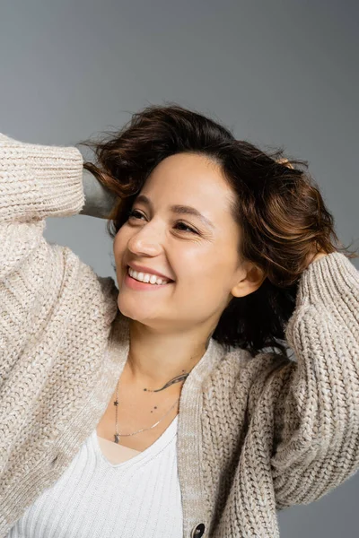 Donna sorridente in cardigan caldo toccando i capelli ondulati e distogliendo lo sguardo isolato sul grigio — Foto stock