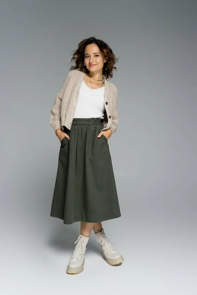 Pleine longueur de jeune femme dans un cardigan confortable et bottes posant avec les mains dans des poches de jupe verte sur fond gris — Photo de stock