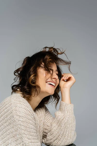 Mujer con el pelo ondulado usando cárdigan caliente y sonriendo aislado en gris - foto de stock