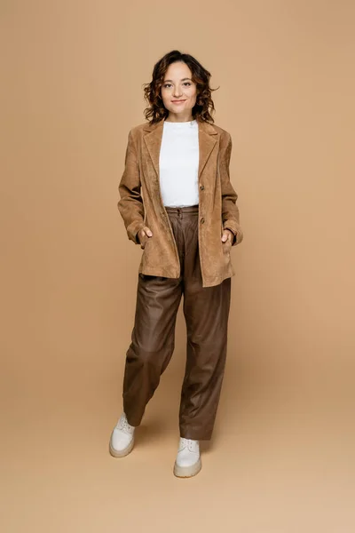 Pleine longueur de femme brune debout avec les mains dans des poches de veste en daim brun sur fond beige — Photo de stock