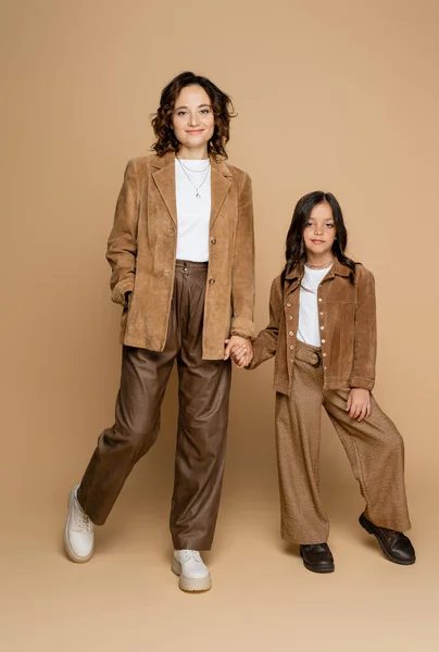 Pleine longueur de maman et fille en pantalon marron et vestes en daim tenant la main sur fond beige — Photo de stock