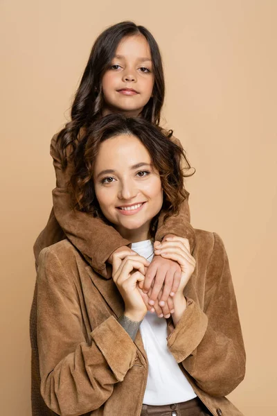 Mujer sonriente en chaqueta de gamuza marrón cogida de la mano de la hija y mirando a la cámara aislada en beige - foto de stock