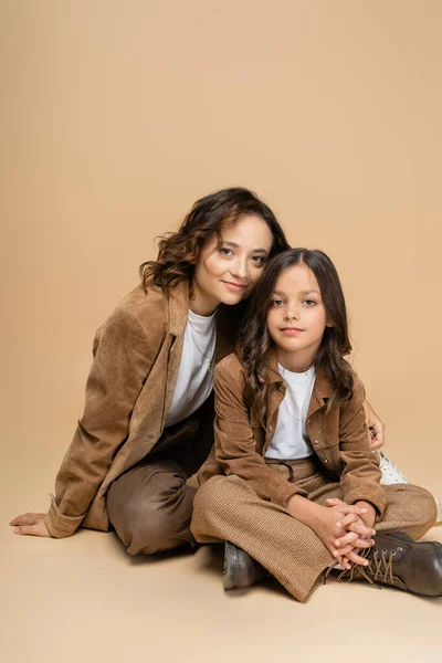 Madre e hija en chaquetas de gamuza y pantalones marrones sonriendo a la cámara mientras están sentadas sobre fondo beige - foto de stock