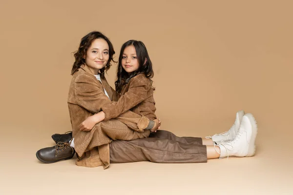 Happy woman and girl embracing while sitting in suede jackets and boots on beige background — Stock Photo