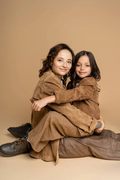 Mother and daughter in suede jackets and brown pants smiling at camera while sitting on beige background — Stock Photo