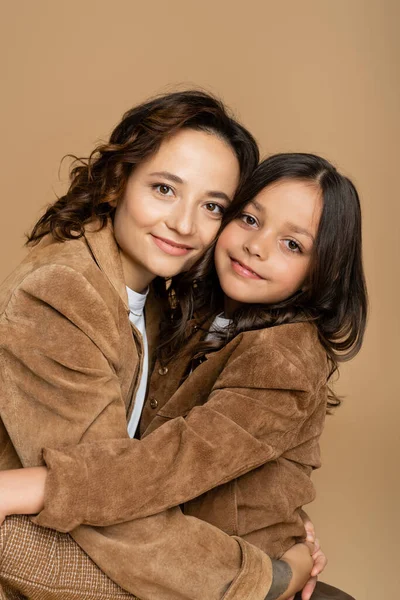 Brunette mother and daughter in brown jackets hugging and smiling at camera isolated on beige — Stock Photo