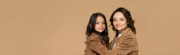 Brunette woman and girl in trendy autumn jackets embracing while looking at camera isolated on beige, banner — Stock Photo
