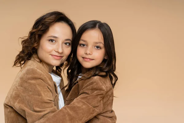 Mamá y su hija en chaquetas de gamuza con estilo sonriendo a la cámara mientras se abraza aislado en beige - foto de stock