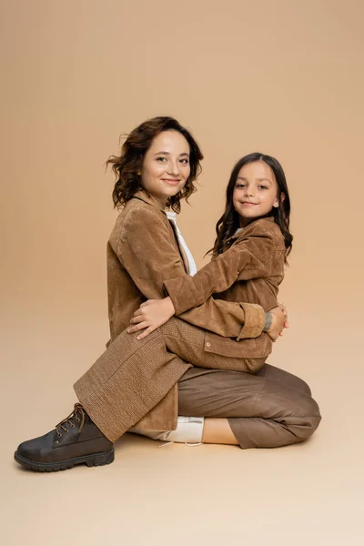 Brunette mother and daughter in fashionable clothes hugging and smiling at camera while sitting on beige background — Stock Photo