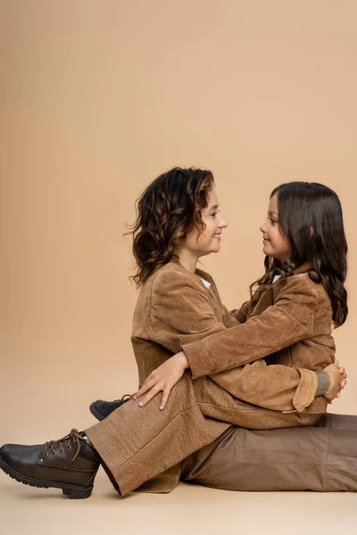 Vista lateral de la mamá y la hija en elegante traje de otoño abrazándose y sonriendo el uno al otro sobre fondo beige - foto de stock