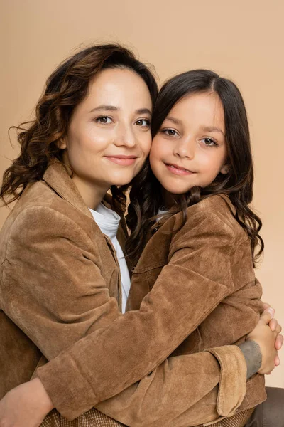 Morena mãe e filha em casacos de outono marrom abraçando e sorrindo isolado no bege — Fotografia de Stock